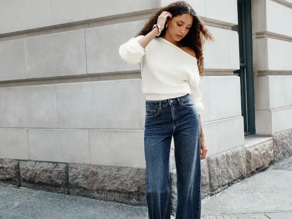 Model is wearing a white off the shoulder sweater with dark wash jeans.